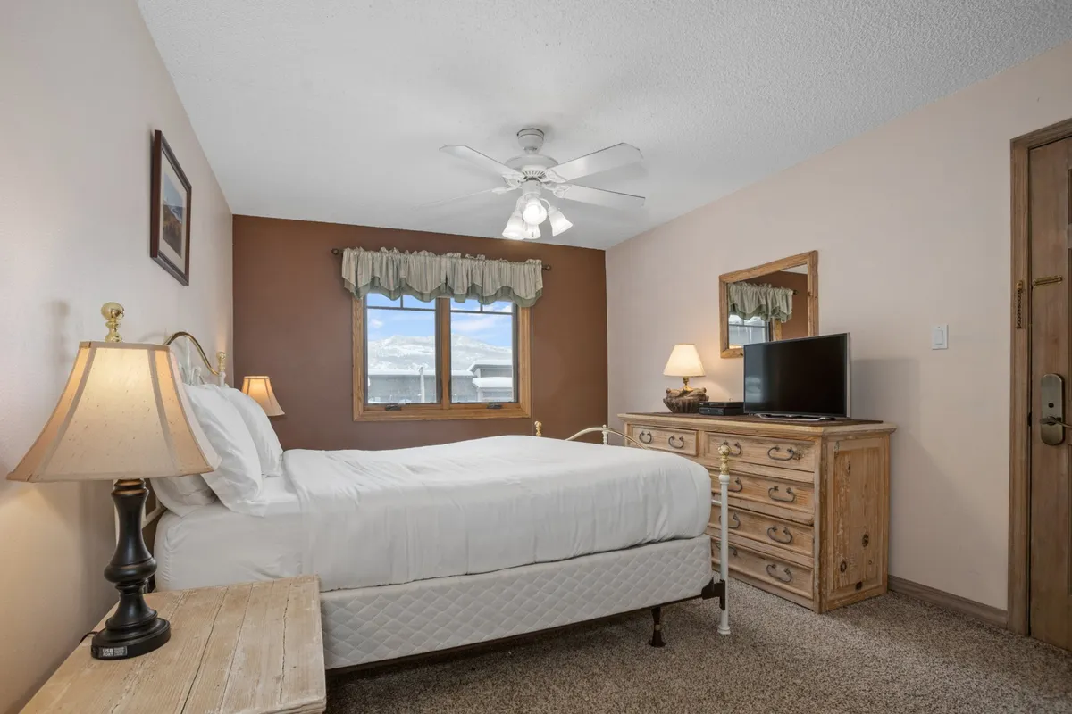 A calm, peaceful bedroom with a bed dressed in white linens, wooden dresser, bedside lamps, and a window offering natural light and a scenic view. The space feels serene, ideal for restful nights and quiet moments.