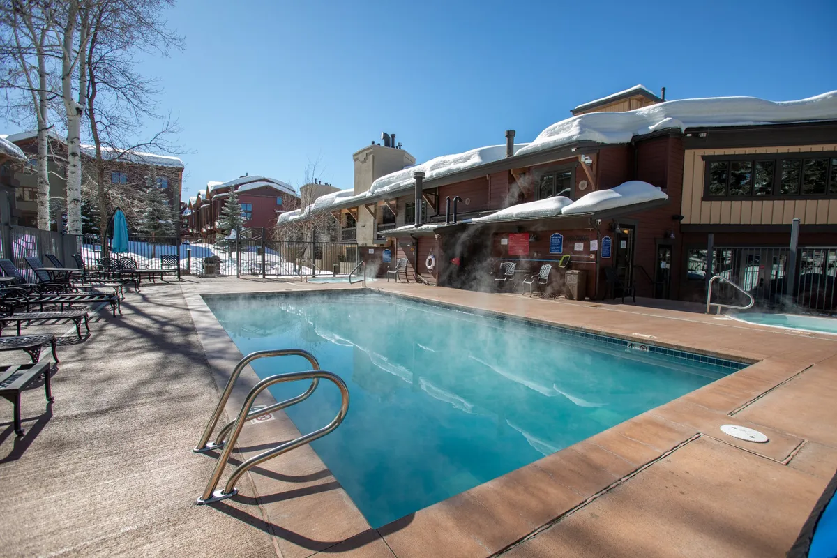 An inviting heated outdoor pool is surrounded by pristine snow and lounge chairs, with the blue sky overhead. The scene highlights the appeal of unwinding in a warm pool after a day on the slopes, with the contrast of the snow creating a truly resort-style, alpine atmosphere.