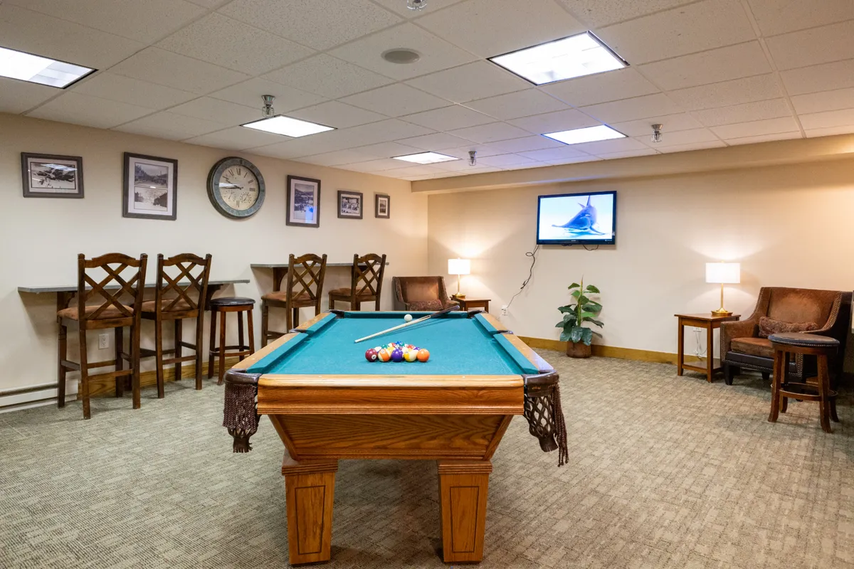 A modern clubhouse lounge with a pool table as the centerpiece, surrounded by bar stools, a cozy seating area, a TV, and stylish wall decor. The layout invites guests to unwind, play games, and connect in a comfortable, social setting.