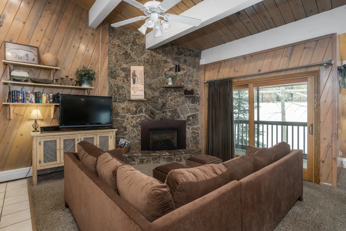 This inviting living area features a stone fireplace, a plush sectional sofa, a large window with mountain views, and a TV strategically placed for movie nights or catching up on the big game. The space is arranged for comfort and togetherness, with ample seating and a welcoming ambiance.
