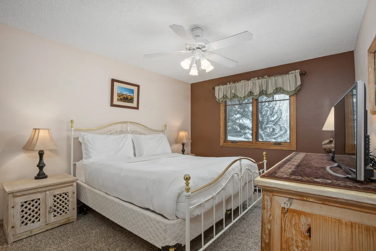A cozy bedroom with a bed featuring crisp white linens, wooden nightstands, a window offering a snowy mountain view, a framed picture on the wall, and a television on top of a dresser. The overall ambiance is warm and inviting, ideal for relaxation after a day of adventure.