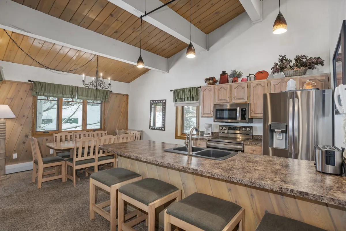A view of the kitchen and dining area highlights a generous kitchen island, a large wooden dining table, exposed wooden beams, stainless steel appliances, and expansive windows that flood the space with mountain light. The setup is ideal for communal cooking and meals, reinforcing the theme of togetherness.
