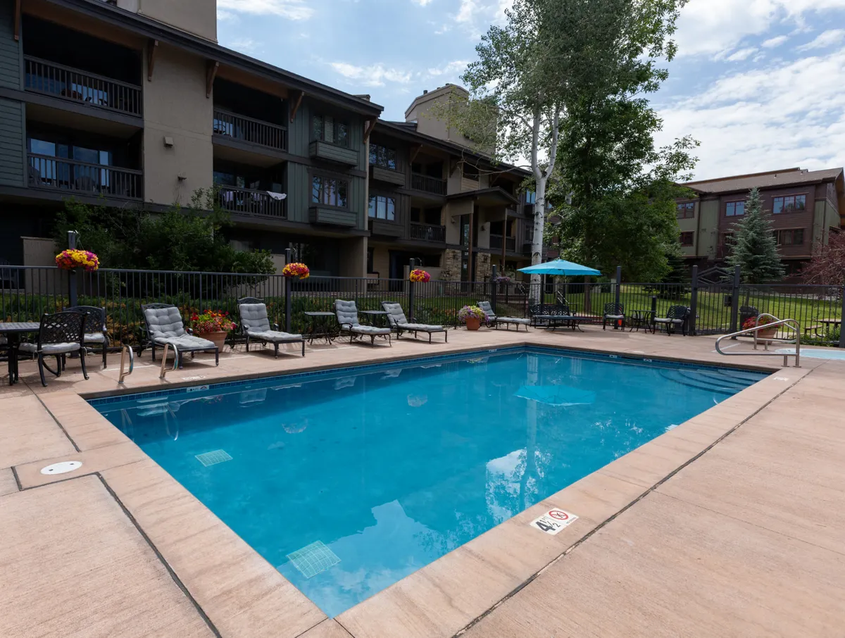 This exterior image features a sunny swimming pool surrounded by comfortable lounge chairs and greenery. The balconies of the apartments overlook the pool, and groups of people are shown swimming, sunbathing, and socializing, creating a lively, family-friendly atmosphere ideal for après-ski relaxation.