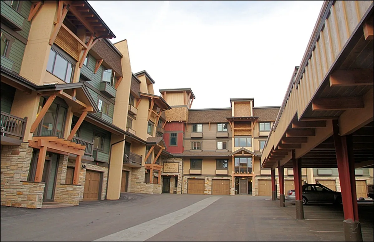 The exterior of the building features wooden beams, a stone facade, colorful accents, and large windows. The setting is welcoming and provides context for the property's mountain location and easy access to the slopes, trails, and shuttle.