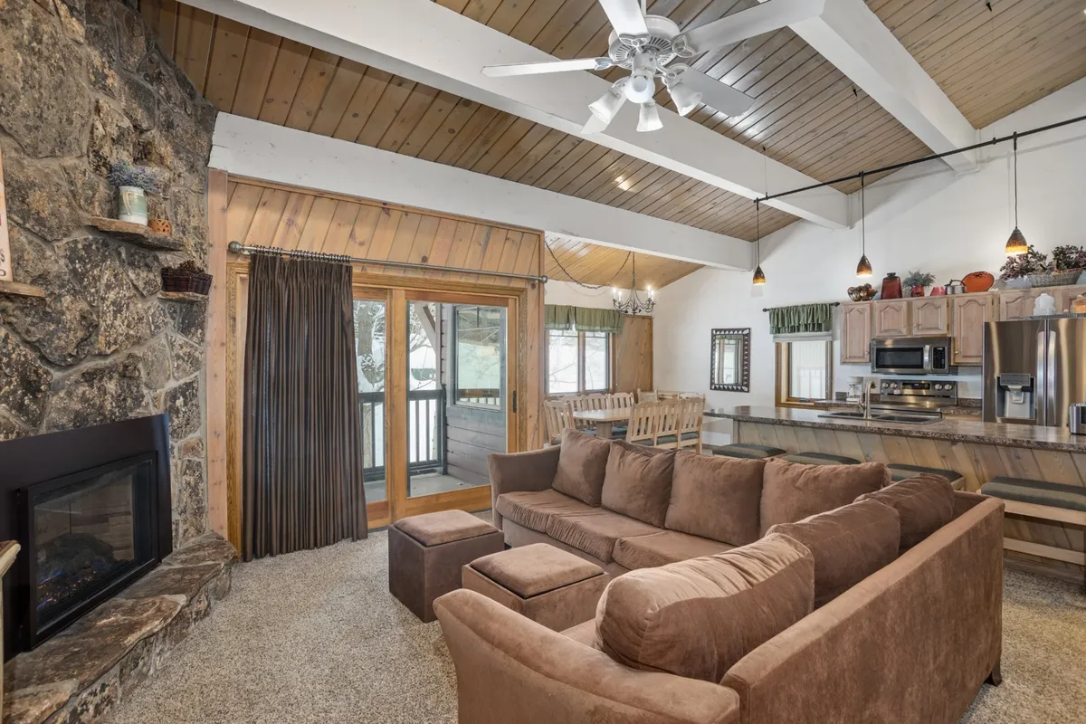 This interior shot captures a spacious living area featuring a striking stone fireplace, a large sectional sofa perfect for group relaxation, a wooden dining table that comfortably seats many, and a glimpse of the connected kitchen area. The room is accented with warm wood tones and receives ample natural light from large windows, highlighting the open-concept layout.