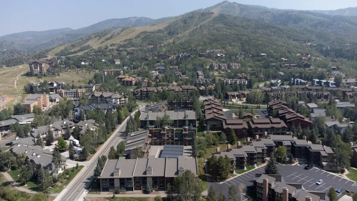 The image features breathtaking mountain vistas towering behind charming condo residences, with a lively open space where people are enjoying outdoor leisure activities. The scene is vibrant, basked in sunlight, and conveys a sense of community and adventure right outside the doorstep.