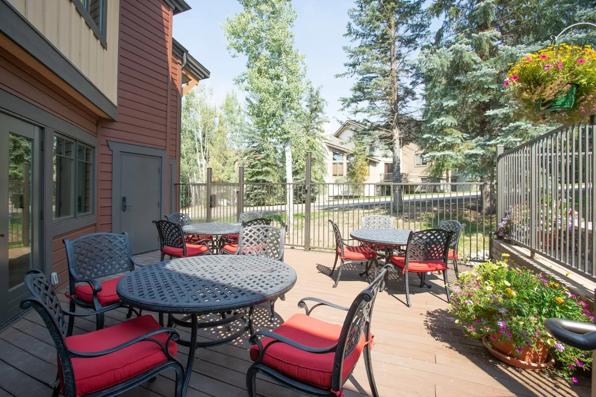 A welcoming outdoor dining area featuring round tables with red cushioned chairs, lush greenery, and a fence. The setup is ideal for social gatherings, morning coffee, or sunset dining surrounded by nature.