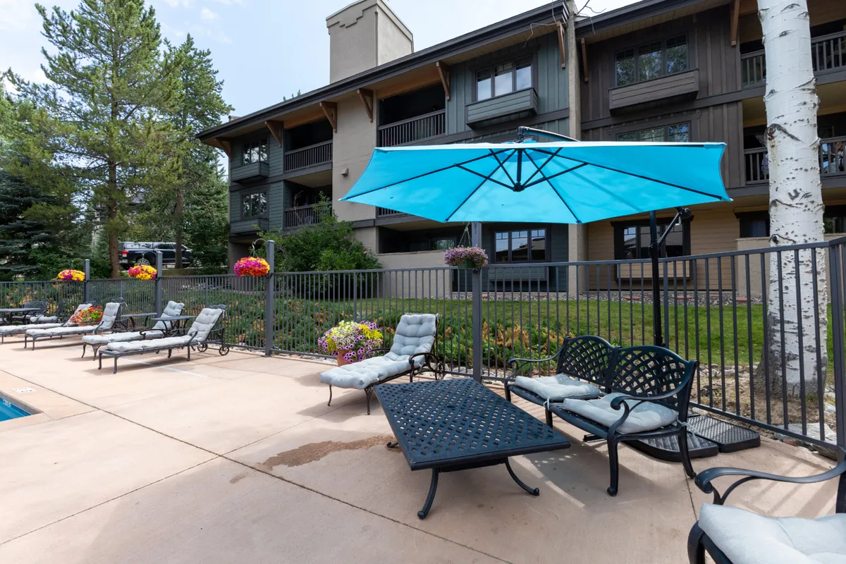 A vibrant outdoor swimming pool area with blue umbrellas, lounge chairs, colorful flower beds, and the building facade visible in the background. The space is designed for leisure, socializing, and group relaxation against a backdrop of natural beauty.