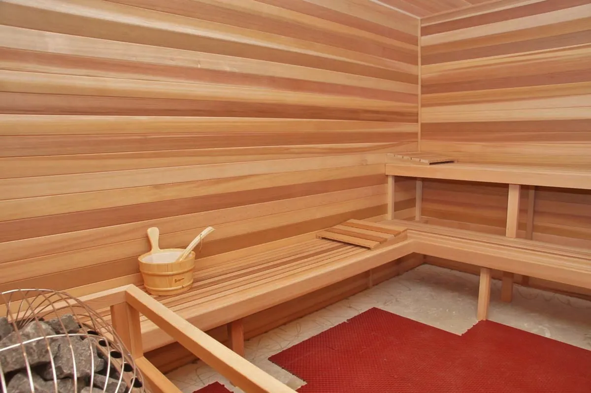 A tranquil sauna room with wooden benches, a central stone heater, a wooden bucket, and a red mat. The space is designed for wellness rituals, relaxation, and quiet reflection, evoking a spa-like retreat within the property.